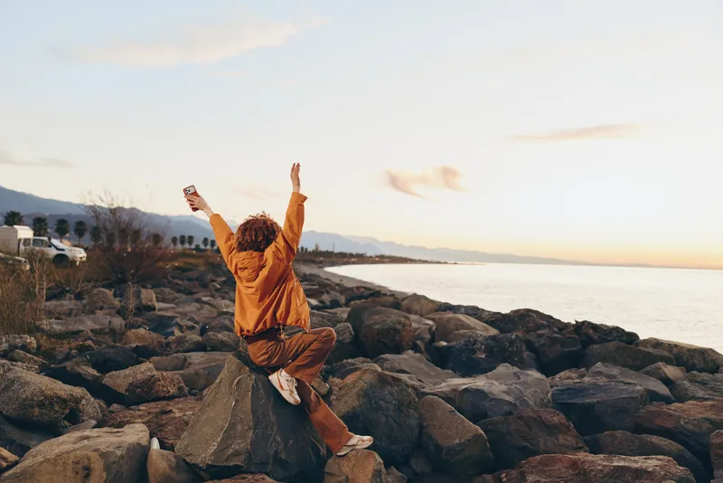 woman lifestyle on a seaside rocky coast, arms raised while jumping on rocks at sunset, warm light and ocean horizon, happiness in outdoors coastal leisure, casual travel portrait