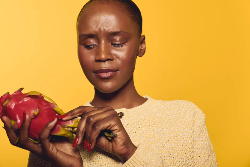 Young black woman with dark skin examines red dragon fruit in hands, wearing beige sweater. Yellow background. Concept of healthy lifestyle, exotic fruit, and natural nutrition.