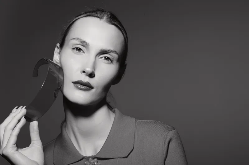 A pale white woman with light skin holds a broken mask near her face, posing in a studio with neutral lighting. Concept for identity, emotion, and vulnerability.