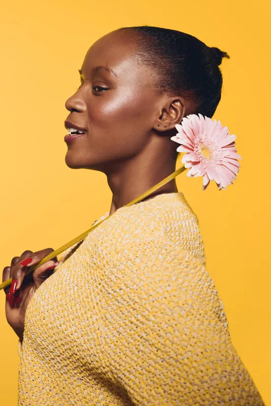 Profile of a smiling black woman with dark skin holding a pink gerbera flower behind her shoulder on yellow background. Concept of beauty, nature, and happiness with floral element.
