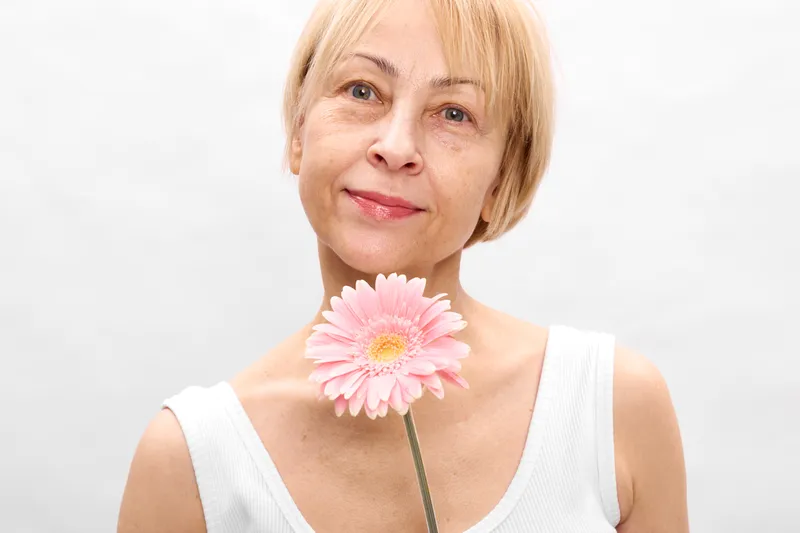 Older woman portrait, pink gerbera flower in hand, soft smile and calm eyes, white studio background, close up headshot, gentle skincare look, spring beauty concept for wellness
