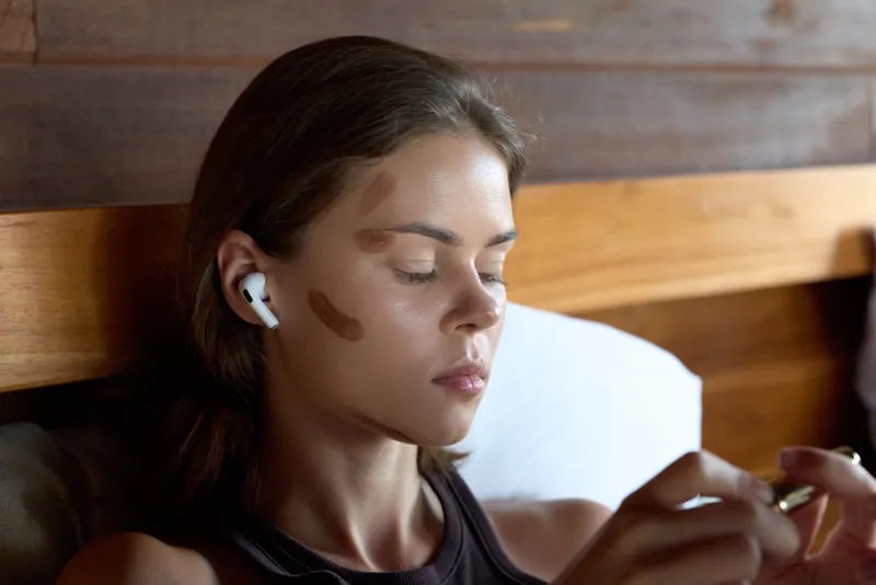 Calm woman with makeup resting against a wooden headboard, wearing a wireless earbud, focused on self care, soft natural light, relaxed lifestyle moment, intimate portrait