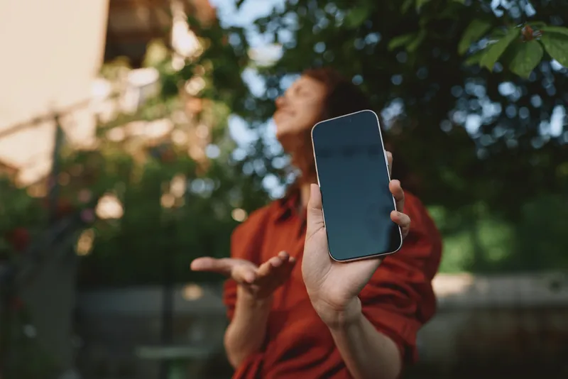 Woman lifestyle outdoors, smiling while holding a phone toward the camera, sunlit moment among greenery, relaxed posture and candid charm in natural colors