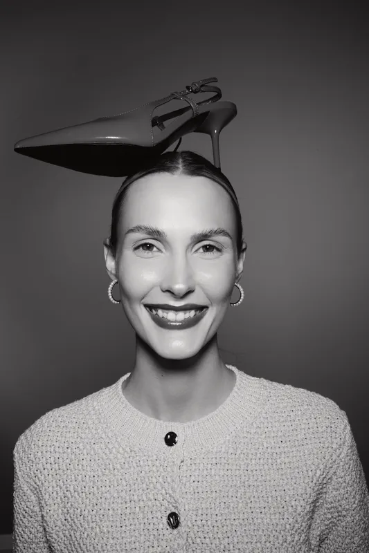 Young white woman with pale skin smiles confidently wearing a knitted sweater and balancing high heel shoe on her head. Studio portrait in black and white with minimalist style.