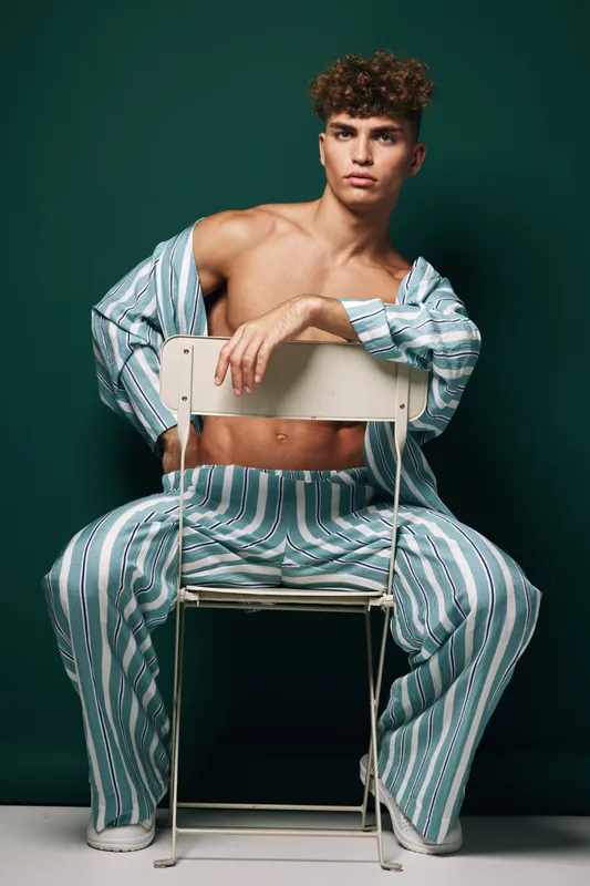 Young man with curly hair wearing striped pajamas sitting on chair backwards with serious expression against dark green background in studio setting.
