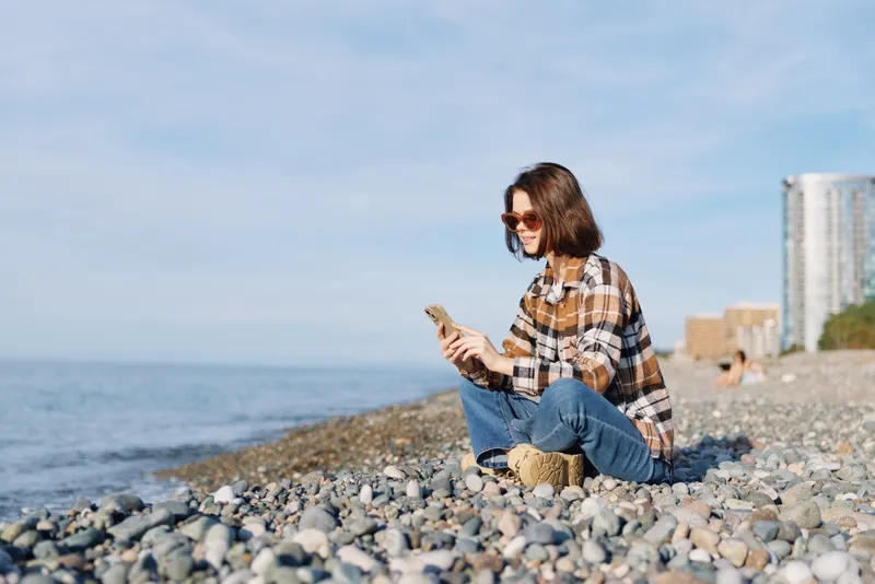 Woman sits on a pebble beach by the sea, wearing a plaid shirt and jeans, looking at her phone with distant buildings, clear blue sky and a light breeze in bright sunlight.