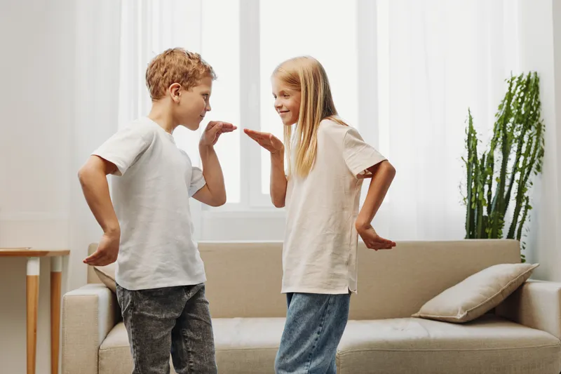 Two smiling children play a hand game in a bright living room with casual clothes and natural light, expressing joy and friendship during a playful moment.