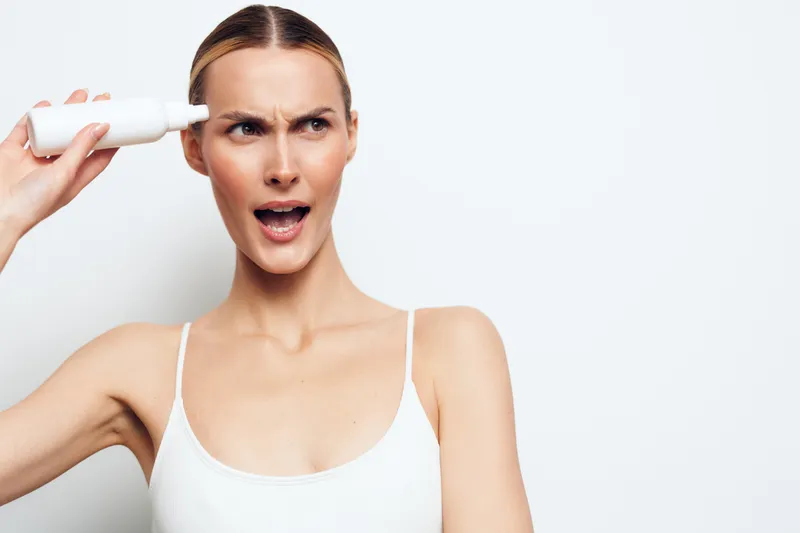 Young woman with clear skin holding a white bottle near her temple with confused expression. White background, casual outfit, concept of skincare and personal care routine.
