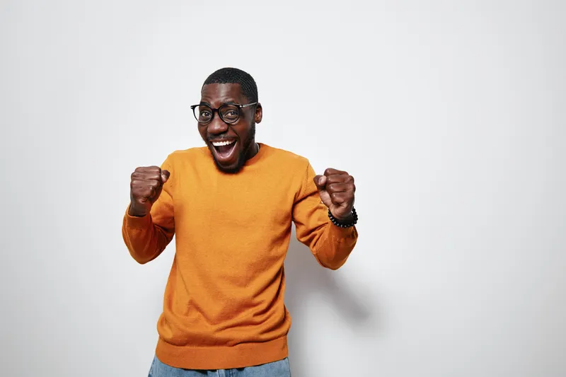Excited black man with dark skin wearing glasses and orange sweater celebrating success with clenched fists and joyful expression on white background in studio.