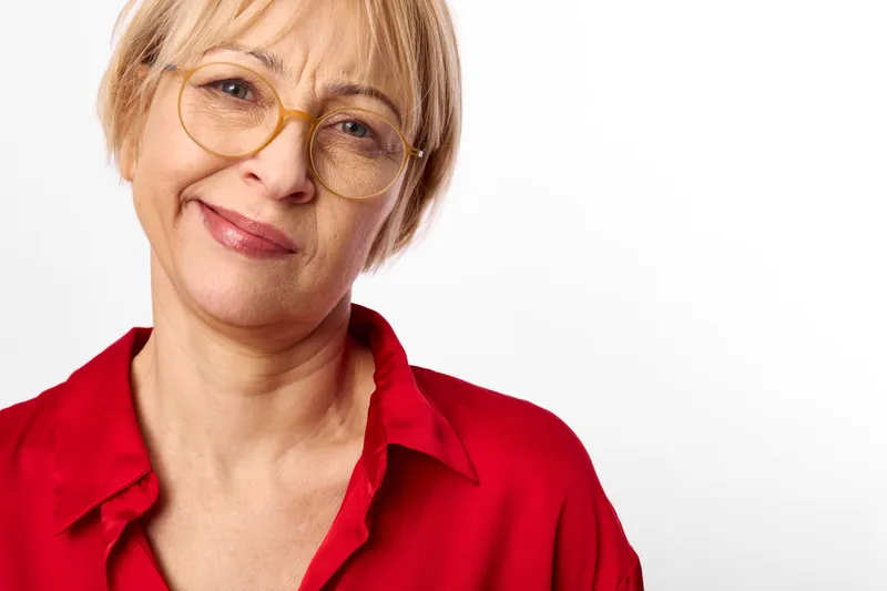 Senior woman portrait, wearing glasses and red shirt, smiling warmly at camera, closeup face, soft studio lighting, clean white background, calm friendly mood, natural expression