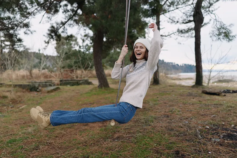 Woman on swing outdoors, smiling and swinging over a grassy lakeside area with trees in cool weather, showcasing carefree joy and natural scenery.