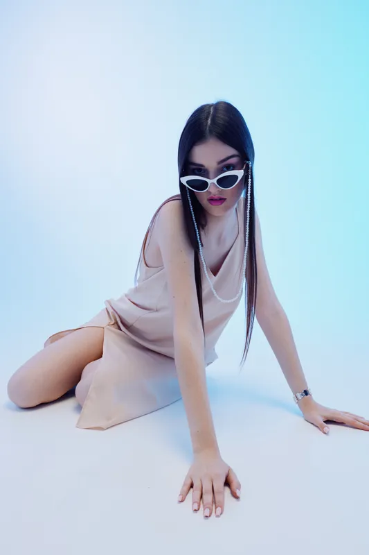 Young woman in a beige dress poses on the floor wearing white sunglasses. Her long hair is styled with beads. Studio shot with soft lighting. Concept of beauty fashion style.