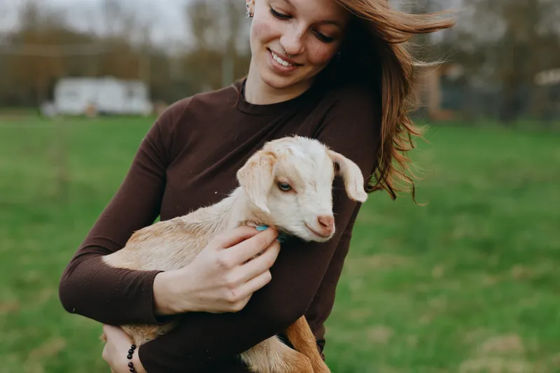 Smiling woman hugs a goat kid in a grassy field, highlighting a gentle countryside lifestyle, connection with animals, and pastoral calm under soft natural light, creating a warm, hopeful mood.
