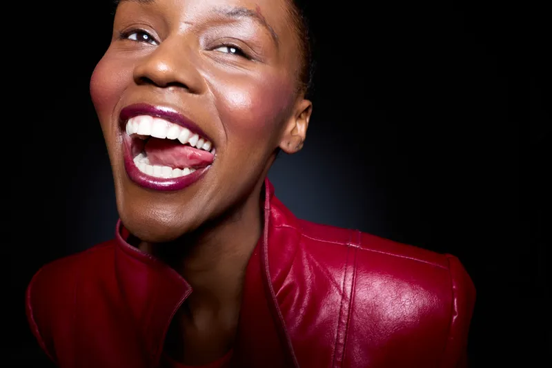 Close-up of a joyful African woman with dark skin laughing widely wearing a red leather jacket. Black background emphasizes her happy expression and vibrant mood. Concept of happiness and confidence.