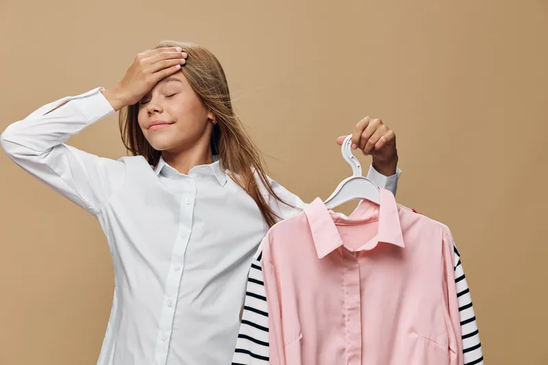 Young woman holding pink shirt on hanger and touching forehead with eyes closed for decision making or frustration in clothing choice indoors with neutral background.