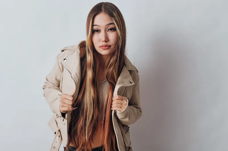 Young woman with long brown hair wearing beige jacket posing confidently against white background. Casual fashion style with natural makeup and relaxed expression in studio setting.