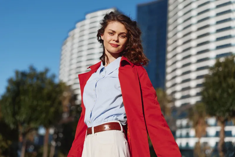 Confident woman outdoors in a red coat, white shirt, and belt, posing in an urban setting on a sunny day, fashion portrait with modern city backdrop and daylight.
