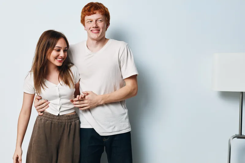 Young couple smiling and embracing while standing against white background. Casual clothes, happiness and affection with man and woman showing friendship and love together.
