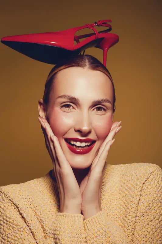 Smiling woman with red high heel shoe on head poses with hands near face against brown background. Concept of fashion, fun, and playful style with bright makeup and cozy sweater.