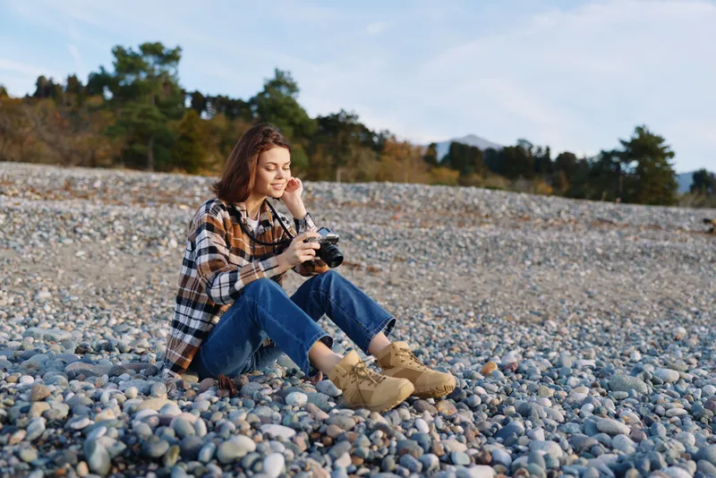 Outdoor woman photographer sitting on a rocky beach with a camera, wearing a plaid shirt and boots, enjoying a peaceful moment among pebbles and distant trees under a clear sky.