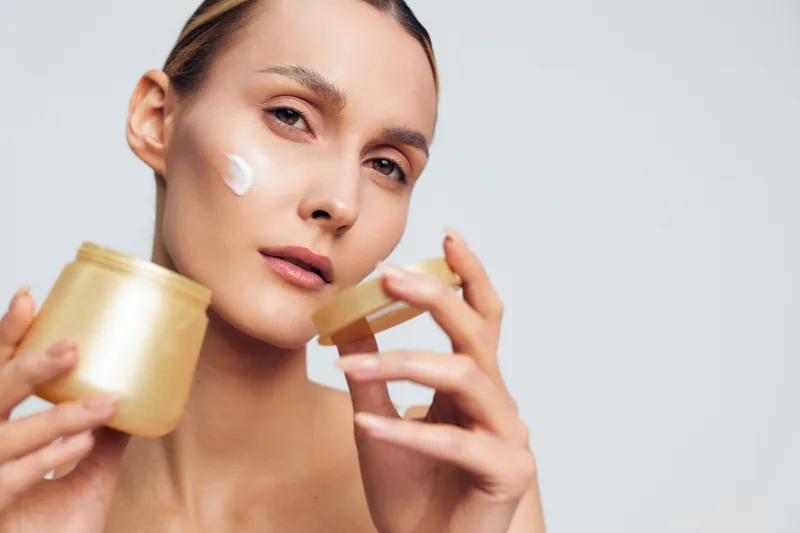 Woman applying face cream with finger, holding a jar, skincare self care concept for wellness, close up on light gray background, soft studio lighting, beauty spa mood