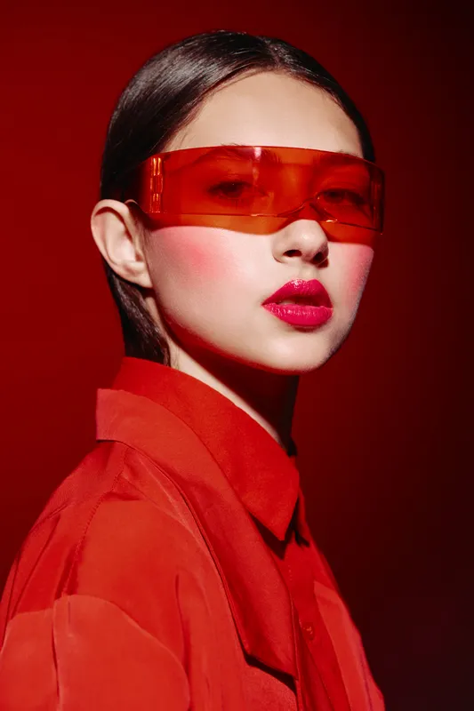 Young woman in bright red outfit and red translucent glasses poses dramatically on red background. Bold fashion and creative beauty concept with striking makeup and styling.