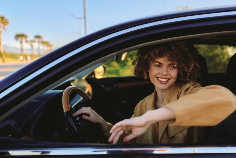 Woman driver sits inside a car with a bright smile, enjoying a casual lifestyle, captured in retro film color on a sunny street with palm trees