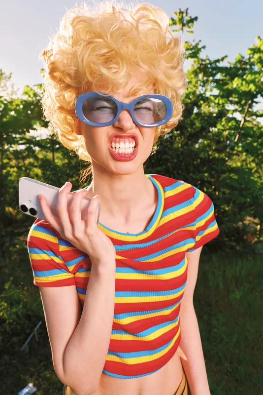 A young woman with a blonde wig and oversized blue glasses poses playfully outdoors, holding a smartphone as she strikes a bold retro expression.