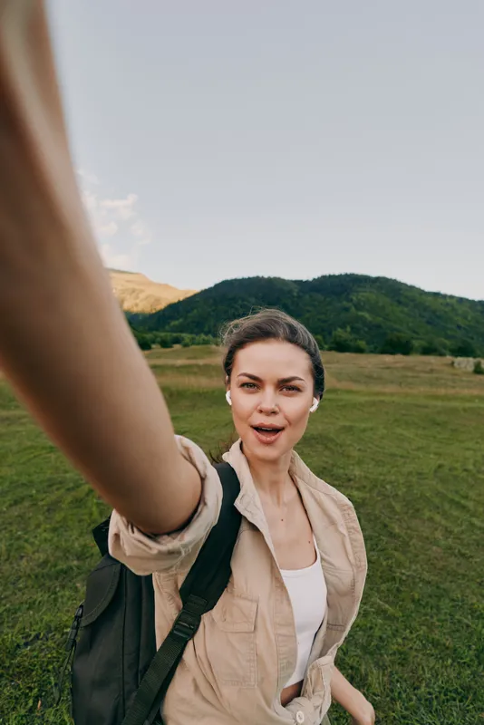 Young woman outdoors taking selfie, casual outfit, backpack on shoulder, green field and hills in background, natural light, relaxed and confident expression