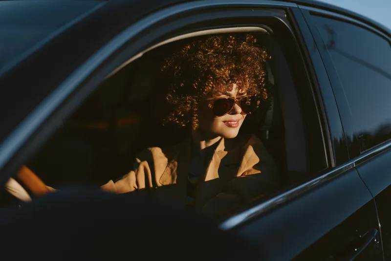 Lifestyle woman drives a car with retro film color, wearing sunglasses and curly hair, warm golden light on her face, confident mood, urban roadside scene. Warm tones evoke film mood.
