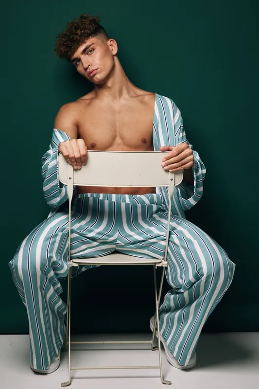 Young man with curly hair wearing striped pajamas sitting backwards on white chair against dark background. Casual pose, relaxed mood and fashion style in studio portrait.