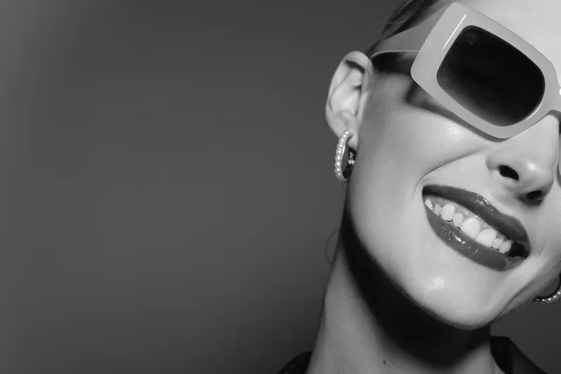 Close-up of a smiling white woman with pale skin wearing sunglasses and earrings. Black and white portrait with stylish confident mood.