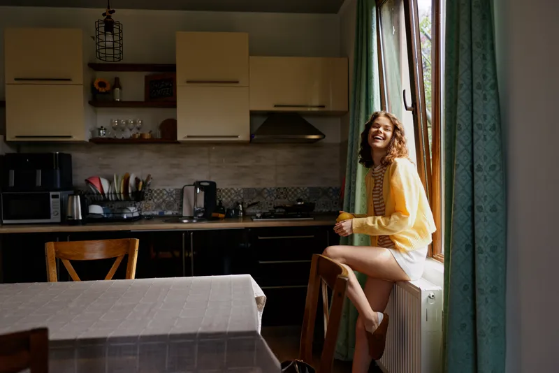 Cozy young woman sits on a radiator by a sunlit kitchen window, wearing a yellow sweater and light shorts, enjoying a calm moment with a soft smile, warmth and home comfort