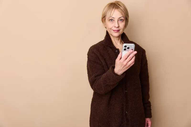 Mature woman portrait holding smartphone, taking selfie, calm confident expression, beige studio background, cozy warm sweater, modern technology for communication concept