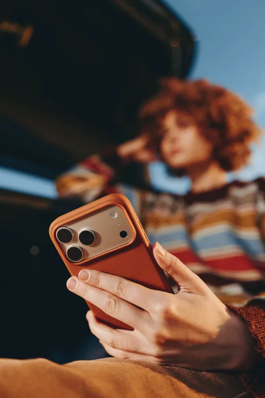 Woman lifestyle outdoors, closeup of hands holding smartphone with orange case, casual striped sweater, blurred curly hair background, sunlit candid moment, warm natural light, soft tones