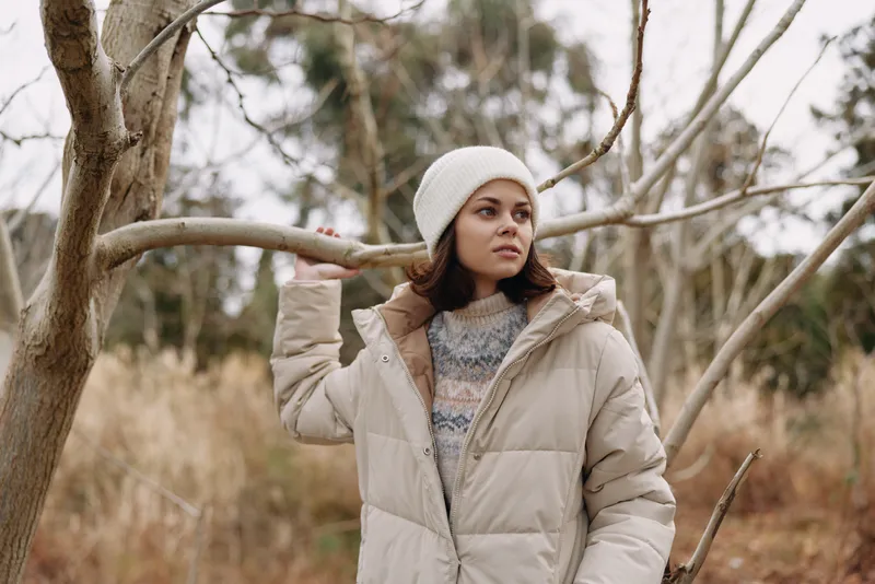 Woman in a light beige puffer coat and knit beanie stands among bare trees in a chilly landscape, posing thoughtfully as autumn hues fade.
