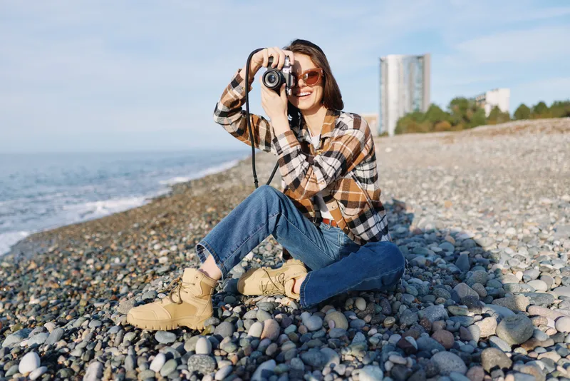 Female photographer sits on a pebbled beach, wearing a plaid shirt and jeans, taking a photo with a camera as waves and distant buildings create a scenic backdrop.