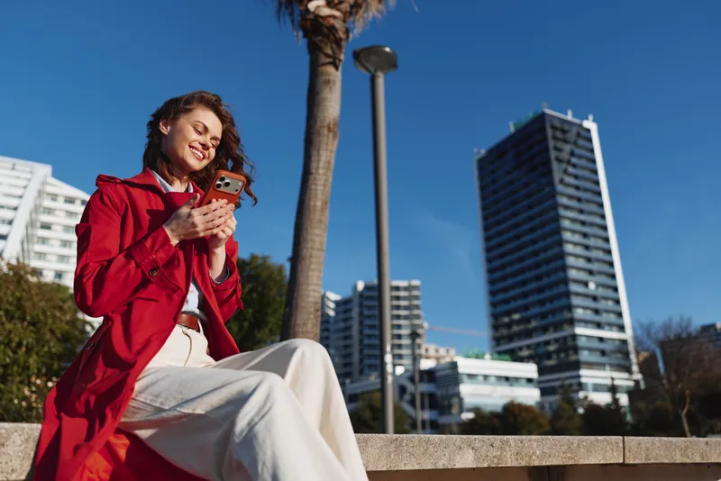 Young woman in a red coat sits on a low wall in a sunny urban park, smiling at her smartphone with relaxed posture as palm trees sway and modern skyscrapers loom behind.