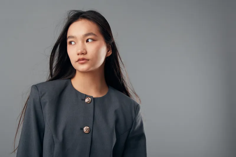 Young woman with long dark hair wearing gray jacket looking away with serious expression on gray background. Portrait of thoughtful female model in simple elegant outfit with natural makeup.