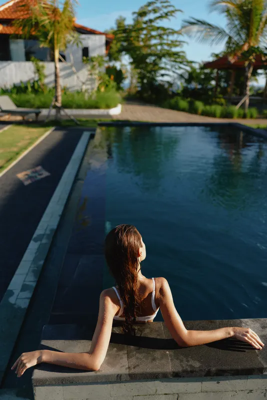 A woman sits at the pool edge with her back to the camera, basking in warm sunlight. Tropical palm trees frame the bright outdoor scene, conveying a lifestyle moment of relaxation, vacation, and