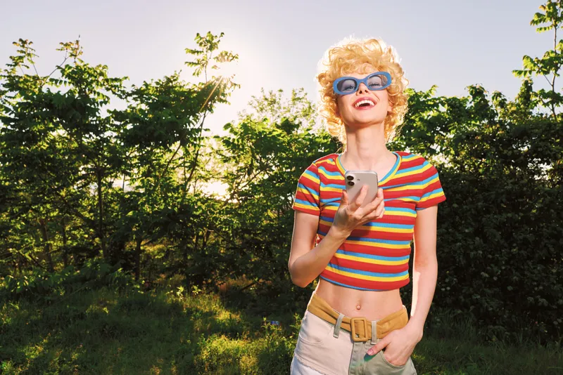Cheerful woman outdoors, wearing a striped shirt and sunglasses, enjoying a sunlit moment with a carefree smile, casual lifestyle vibe and retro film color palette. bright tones here