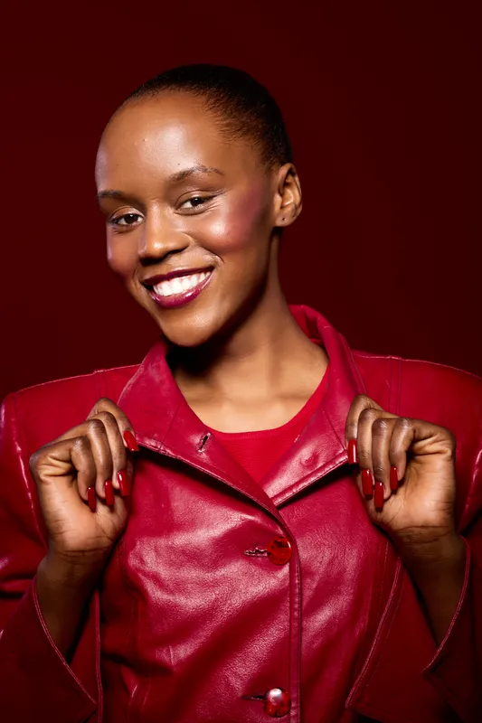 Smiling African woman with dark skin wearing red leather jacket poses confidently against red background. Concept of fashion, beauty, confidence, and vibrant style for dark skin tone.