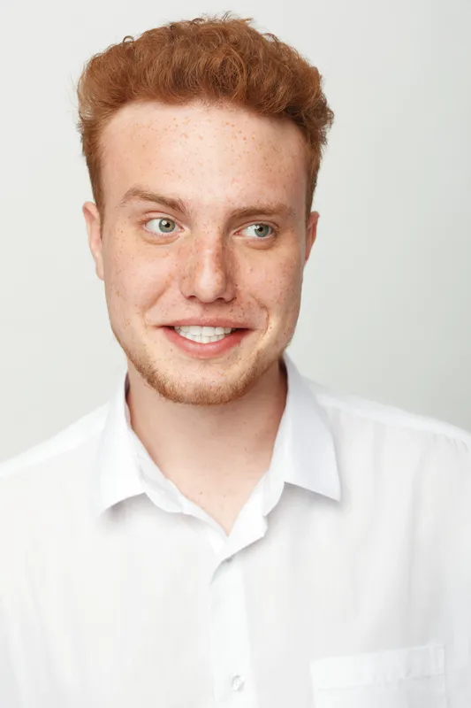 Smiling young man with red hair and freckles wears white shirt, looking aside on light background. Portrait of casual confident male with natural expression and clean style.