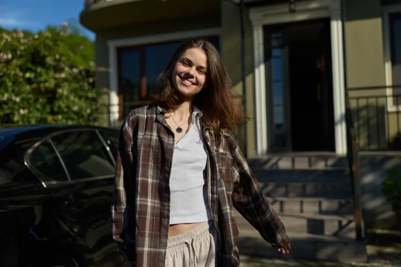 Candid portrait of a smiling woman in plaid shirt and light top, standing outdoors in a sunny residential area, conveying casual lifestyle and relaxed mood today