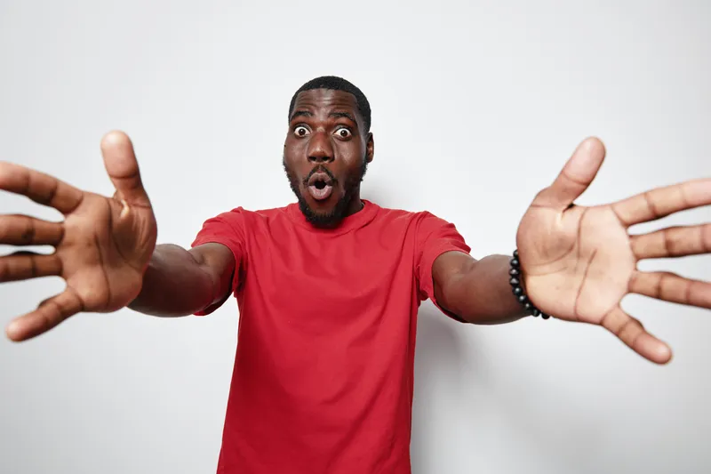 African American man with dark skin and beard wearing red t shirt with surprised expression reaching hands forward isolated on white background for emotion and reaction.