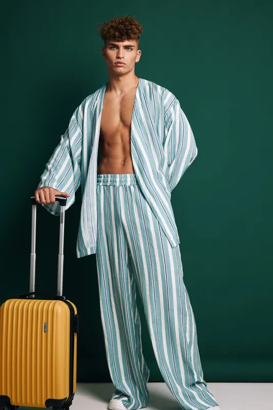Young man in striped pajamas standing with yellow suitcase on dark green background, casual travel outfit and relaxed pose for vacation or trip concept.