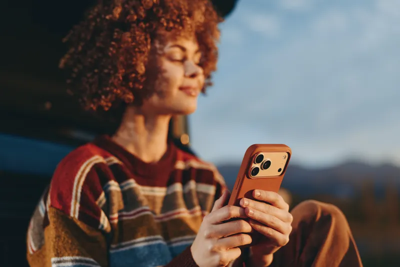 Woman with curly hair smiling while holding smartphone outdoors in evening light, casual plaid sweater, relaxed lifestyle mood, soft focus mountains background, communication concept