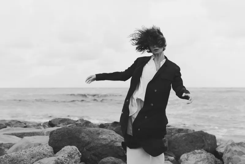 Windswept figure stands on a rocky shore, blazer fluttering, wind blown hair, monochrome tones, dramatic coastal fashion mood with sea breeze and moody movement along the coastline