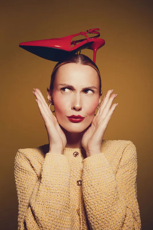 Woman with red high heel shoe balanced on her head looks confused and thoughtful. She wears a beige textured sweater and gold earrings on a warm brown background. Concept of fashion and creativity.