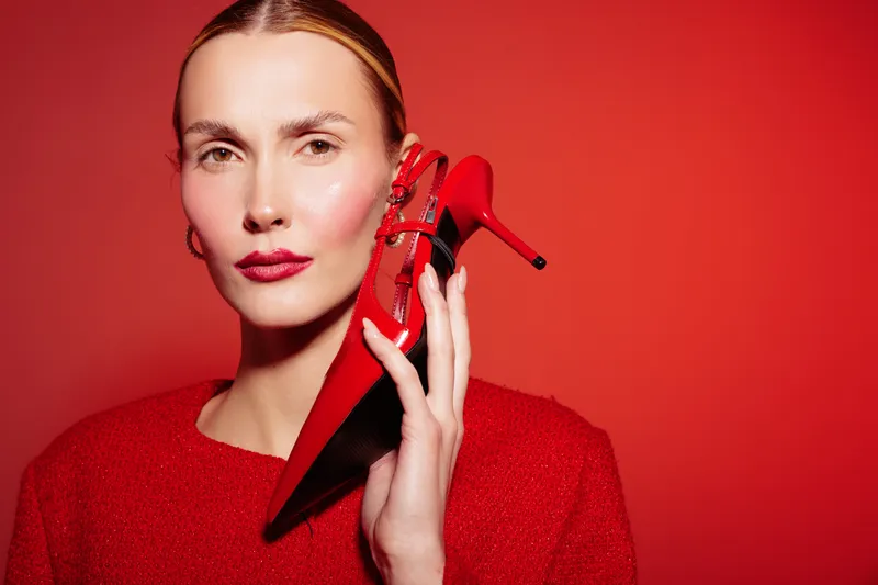 Stylish woman in red sweater holds red high heel shoe near face against red background. Concept of fashion, elegance, beauty, and monochrome style with confident female model in studio lighting.